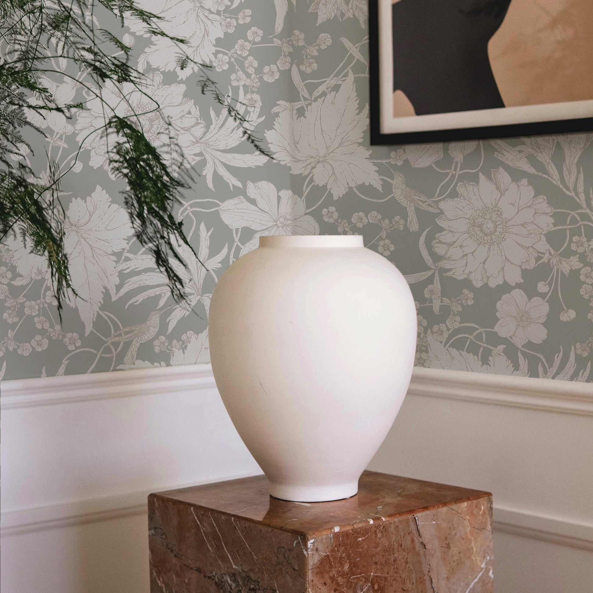 White vase on a wooden pedestal against a floral wallpapered wall.